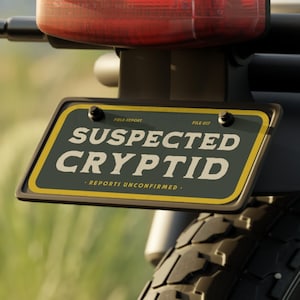 Op de afbeelding: De achterkant van een motorfiets met een rechthoekig rood achterlicht, twee oranje knipperlichten en een zwarte kentekenplaat met een gele rand. De plaat toont de tekst SUSPECTED CRYPTID in dikke witte letters met daaronder REPORT UNCONFIRMED.