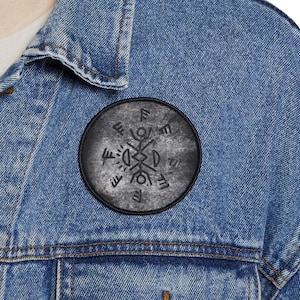 Op de afbeelding: Een ronde, zwarte patch met een wit, gestileerd symbool. De patch is bevestigd op een spijkerjasje. Het symbool is een complex arrangement van lijnen en vormen, mogelijk van historische of culturele betekenis.