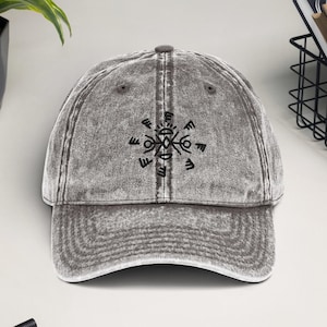 Könnte beinhalten: Eine graue, abgenutzte Baseballkappe aus Denim mit einem schwarzen gestickten Symbol auf der Vorderseite. Das Symbol ist ein Kreis mit einem Stern in der Mitte und acht nach außen zeigenden Pfeilen.