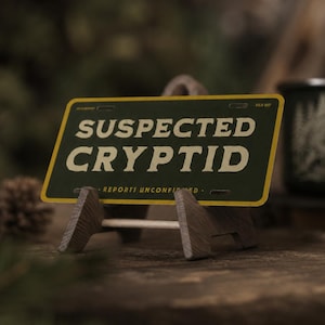 Op de afbeelding: Decoratieve nummerplaat met de tekst "SUSPECTED CRYPTID" in witte letters op een donkergroene achtergrond met een gele rand. De plaat wordt gepresenteerd op een kleine houten ezel. Een donkergroene mok met een bosmotief staat op de achtergrond.