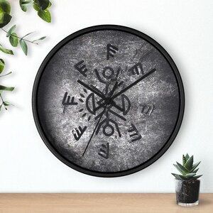 May include: A round wall clock with a black frame and a textured, dark gray, stone-like face. The clock features black hands and a central graphic design consisting of black geometric lines, circular shapes, and runic-style symbols arranged radially.