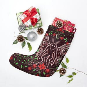 May include: A dark brown Christmas stocking featuring a large moth illustration with white eyes, surrounded by red roses, green vines, and a crescent moon. The stocking is filled with red gift boxes and decorated with pinecones and silver ornaments.