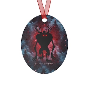 Puede incluir: Adorno ovalado con una cinta roja. El diseño presenta una silueta negra de una criatura con cuernos y ojos rojos brillantes sobre un fondo de bosque y tonos rojos y azules. El texto dice: "hold on to your horns."