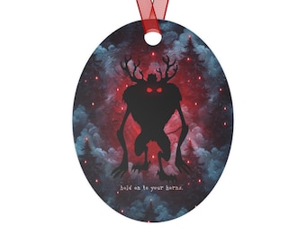 May include: Oval-shaped ornament with a red ribbon. The design features a black silhouette of a creature with antlers and glowing red eyes against a backdrop of a forest and red and blue hues. Text reads: "hold on to your horns."