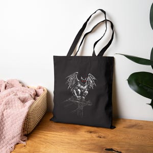 May include: Black tote bag featuring a detailed white illustration of a winged creature with red eyes perched on a utility pole. The bag has black handles and is made of a durable fabric, suitable for carrying various items.