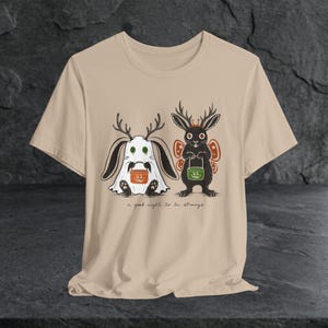 May include: A beige short-sleeve t-shirt featuring a graphic of a ghost-costumed rabbit with antlers holding an orange bucket and a black rabbit with antlers and butterfly wings holding a green bucket, with the text a good night to be strange.