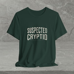 May include: A dark forest green short-sleeved t-shirt featuring the text "SUSPECTED CRYPTID" printed in a light beige, distressed, sans-serif font across the chest, with the smaller text "OBSERVATION STATUS OPEN" positioned directly below it.