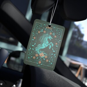 May include: A rectangular car air freshener hanging from a white string, featuring a dark gray background with a teal and peach-colored illustration of a mythical dragon-like creature surrounded by intricate floral vines and a decorative border.