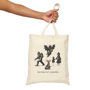 May include: Cream-colored tote bag featuring a black illustration of mythical creatures, including a Bigfoot, a bat-like creature, a deer, and two figures. The text "MOTHER OF LEGENDS" is printed below the illustrations. The bag is held by a person.
