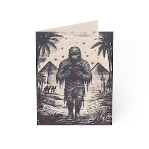 May include: Greeting card featuring a black and white illustration of a mummy holding a red heart. The background includes pyramids, palm trees, and camels. Small red hearts are scattered across the top of the card.