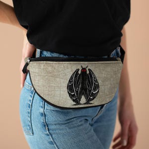 Può includere: Un marsupio beige con un'illustrazione grafica in bianco e nero di una falena con occhi rossi prominenti. La borsa presenta una chiusura a zip nera e una tracolla nera, con un sottile motivo a griglia simile a una mappa sulla superficie.