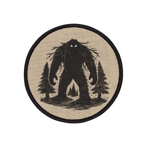 Op de afbeelding: Ronde patch met een zwarte illustratie van een Bigfoot-achtig wezen dat tussen twee dennenbomen staat. Het wezen heeft een ruig, donker silhouet met gloeiende witte ogen. De patch heeft een beige achtergrond en een zwarte rand.