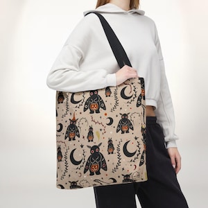 Puede incluir: Un bolso tote de color beige con un patrón repetitivo de criaturas negras similares a polillas con ojos rojos, cada una sosteniendo una pequeña calabaza naranja. El diseño incluye lunas crecientes negras, enredaderas finas y puntos naranjas.