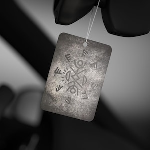 May include: A rectangular car air freshener with a dark, weathered gray stone texture. The surface features a central geometric design composed of stylized stick figures and runic-like symbols arranged in a circular pattern, suspended by a white string.