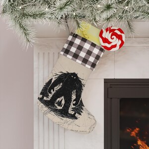 May include: A beige Christmas stocking featuring a black graphic of a yeti standing in a forest. The cuff has a black and white buffalo plaid pattern. The fabric displays a faint map print with text including Greenland and Arctic Circle.