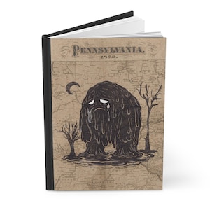 Può includere: Un diario con copertina rigida e dorso nero. La copertina presenta una mappa vintage della Pennsylvania con la scritta "Pennsylvania 1872". Una creatura piangente a forma di blob è illustrata sulla copertina, insieme a una luna crescente e alberi spogli.