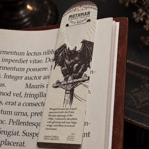 May include: A cream-colored paper bookmark featuring a black ink illustration of the Mothman perched on a utility pole. The text reads Mothman Regional Folklore Field Archive and includes a description of the winged humanoid cryptid with red eyes.