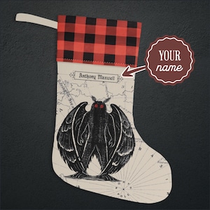 May include: A Christmas stocking featuring a red and black buffalo plaid cuff and a cream-colored body printed with a vintage map background. The center displays a black moth-like creature with glowing red eyes and the text Anthony Maxwell.