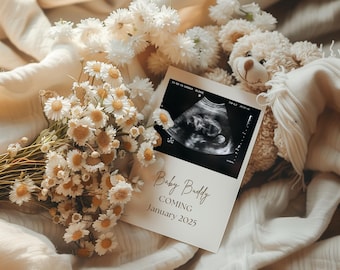 Puede incluir: Una tarjeta blanca con el texto "Baby Buddy Coming January 2023" sobre una manta blanca con un ramo de margaritas secas y un osito de peluche. La tarjeta también incluye una imagen de ultrasonido en blanco y negro.