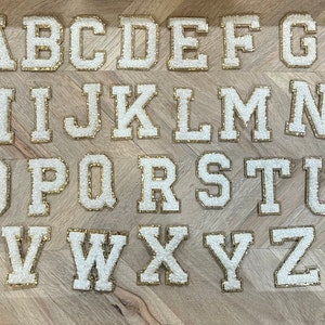 May include: White felt letters of the alphabet with gold trim, arranged in alphabetical order on a wood background.