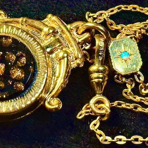 May include: A gold-toned pendant featuring a circular black center with embedded gold flecks, surrounded by a decorative frame. Attached to a gold chain, the necklace includes a small rectangular accent piece with a central iridescent opal stone.