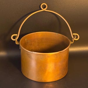 May include: A cylindrical copper pot with a warm metallic finish and a patina. The pot features a curved metal handle attached to two side loops, with a central circular loop at the top of the handle for hanging or carrying the vessel.
