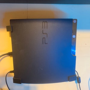 May include: A black PlayStation 3 console with the PlayStation logo on the front. The console is mounted on a wall with wires connected to it.