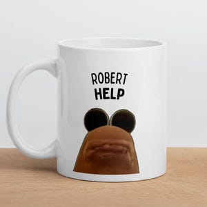 Puede incluir: Taza de cerámica blanca con el texto "ROBERT HELP" en negro. La taza presenta una imagen de dibujos animados de una criatura marrón con grandes ojos negros. La taza está sobre una superficie de madera.