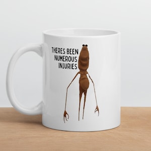 Puede incluir: Taza de cerámica blanca con una figura marrón de dibujos animados y el texto "THERES BEEN NUMEROUS INJURIES" en negro. La figura tiene un cuerpo largo, brazos delgados y una cabeza con dos pequeños ojos negros.