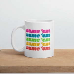 Puede incluir: Taza de cerámica blanca con el texto "name 'em" en un arcoíris de colores. El texto está apilado verticalmente, cada línea en un color diferente: rosa, azul, verde, amarillo y rojo. La taza está sobre una superficie de madera.