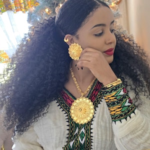 May include: A woman wearing a white and gold traditional Ethiopian dress with a large gold pendant necklace and matching earrings. The necklace and earrings feature a circular design with intricate details.