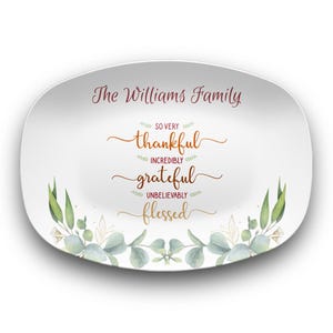Puede incluir: Plato de servir ovalado blanco con un diseño de hojas verdes alrededor del borde. El texto "The Williams Family" está impreso en negro en la parte superior. El texto "So very thankful, Incredibly grateful, Unbelievably blessed" está impreso en rojo y dorado.
