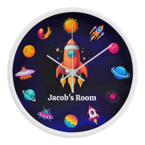 May include: A white round clock with a blue background and a rocket ship in the center. The clock has a white frame and silver hands. The background is decorated with planets, stars, and comets. The text "Jacob's Room" is printed below the rocket ship.