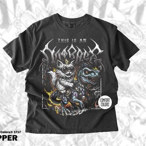 May include: Pepper-colored t-shirt featuring a graphic of a cat with large eyes and a dinosaur-like creature. The text "THIS IS AN" is above the graphic, with the word "SCRAPE" in a stylized font. The shirt is from Comfort Colors.