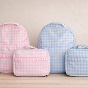May include: A set of four bags featuring a gingham pattern, including two backpacks and two rectangular lunch bags. One backpack and one lunch bag are light pink, while the other backpack and lunch bag are light blue. Each bag has a ruffled trim.