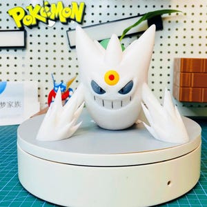 Może przedstawiać: Biała figurka Pokémona wydrukowana w 3D, przedstawiająca Pokemona typu Duch z żółtym kółkiem na czole i ostrymi, kolczastymi odrostami. Figurka stoi na białym talerzu obrotowym.