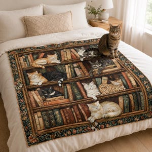 Pode incluir: Uma manta retangular com um design estampado de vários gatos a descansar em estantes de madeira cheias de livros. A manta tem uma borda floral escura e está exposta sobre uma colcha branca com um gato malhado sentado em cima.