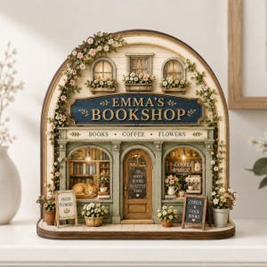 May include: A miniature arched storefront model labeled Emma's Bookshop with a blue sign, green facade, and floral accents. The display features a cat, bookshelves, a coffee machine, and small signs reading Fresh Flowers and Coffee & Good Books.