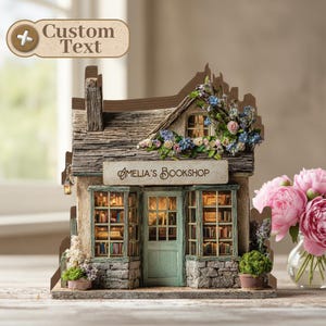 Op de afbeelding: Een gedetailleerd miniatuurmodel van Amelia's Bookshop, met een stenen gevel, een turquoise deur en ramen gevuld met boeken. Het dak is versierd met bloemen. Het bord boven de deur luidt "Amelia's Bookshop."
