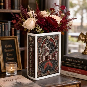 May include: A rectangular white ceramic vase shaped like a book, featuring a detailed relief illustration of Dracula and a woman, the title "DRACULA" in bold lettering, and a dark castle scene. It is filled with cream roses and dark red flowers.