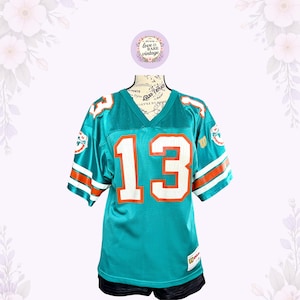 May include: A teal mesh football jersey featuring the number 13 in white with orange outlines on the front and sleeves. The jersey includes white and orange stripes on the sleeves, a V-neck collar, and a Wilson brand logo patch on the lower left.