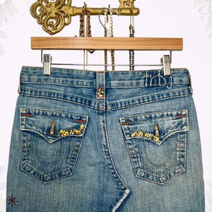 May include: Light blue denim mini skirt with decorative stitching and embellished back pockets. The skirt has a frayed hem and is hanging on a wooden hanger. The back pockets are adorned with yellow and gold embellishments.