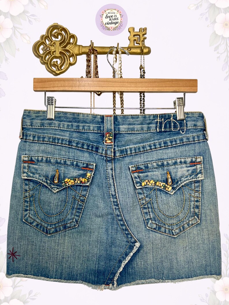 May include: Light blue denim mini skirt with decorative stitching and embellished back pockets. The skirt has a frayed hem and is hanging on a wooden hanger. The back pockets are adorned with yellow and gold embellishments.