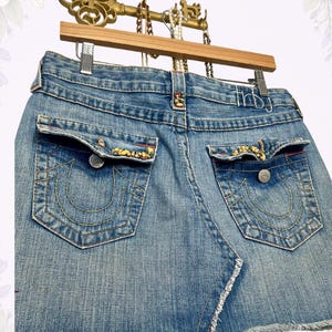 May include: Light blue denim mini skirt with back pockets featuring decorative stitching and embellishments. The skirt has a frayed hem and is hanging on a wooden hanger with a decorative key.