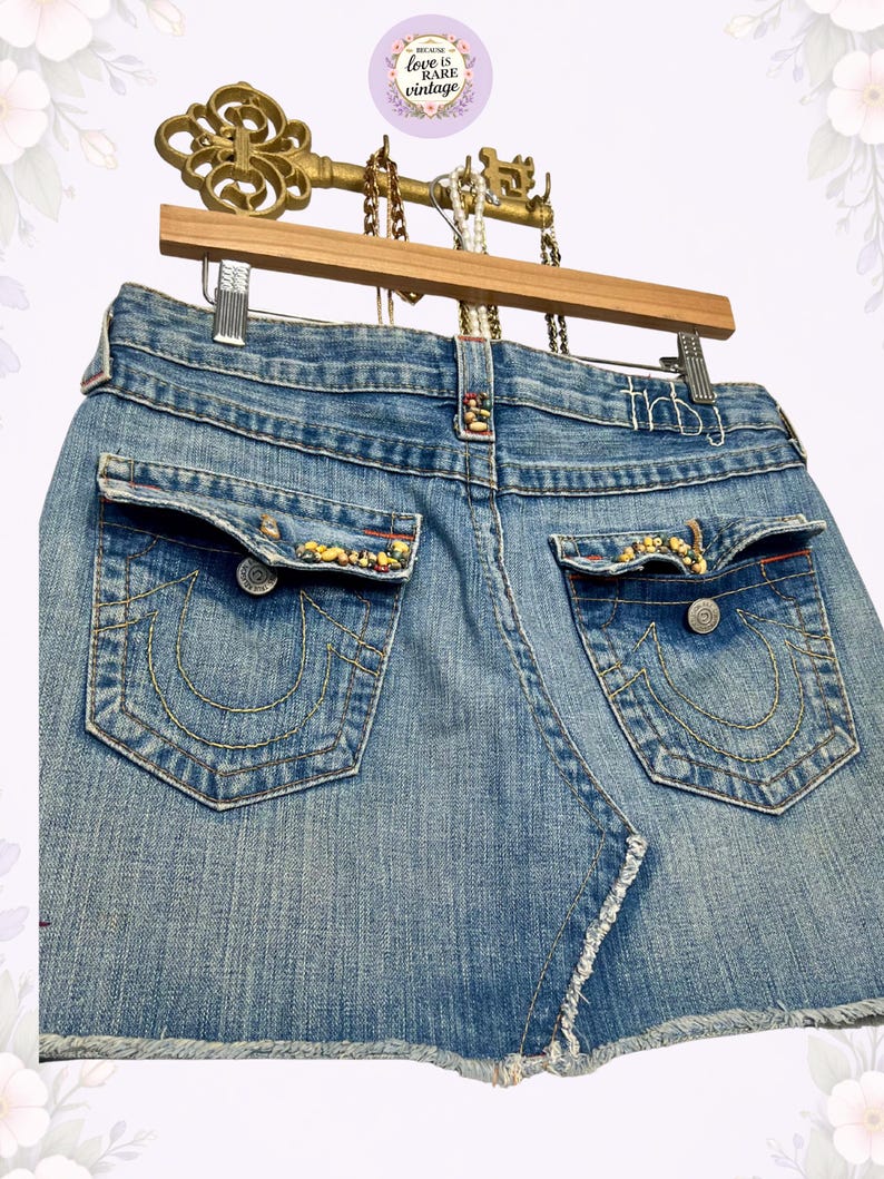 May include: Light blue denim mini skirt with back pockets featuring decorative stitching and embellishments. The skirt has a frayed hem and is hanging on a wooden hanger with a decorative key.