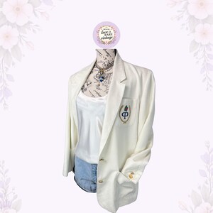 May include: Cream-colored blazer with gold buttons and a pocket featuring an embroidered emblem. The blazer is paired with a white top and denim shorts. A necklace with a blue pendant is visible. The blazer is displayed on a mannequin.