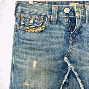 May include: Light blue denim skirt with a distressed look. Features a front zipper, decorative beading on the pocket, and a frayed hem. The skirt has a small embroidered flower detail and orange stitching.