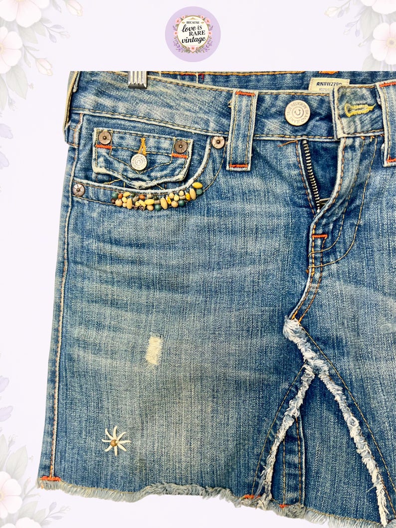 May include: Light blue denim skirt with a distressed look. Features a front zipper, decorative beading on the pocket, and a frayed hem. The skirt has a small embroidered flower detail and orange stitching.