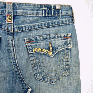 May include: Close-up of a light blue denim skirt with a frayed hem. The back pocket is embellished with colorful beads and gold stitching. The word "thbj" is stitched on the waistband. The skirt is vintage.