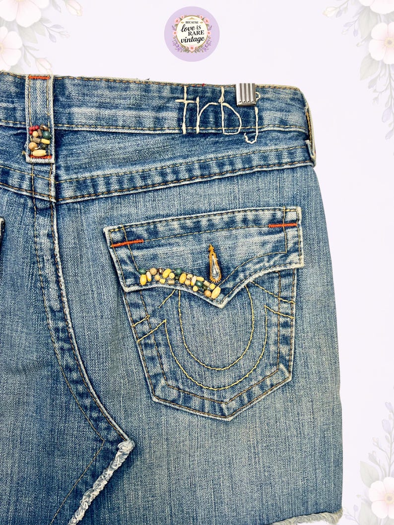 May include: Close-up of a light blue denim skirt with a frayed hem. The back pocket is embellished with colorful beads and gold stitching. The word "thbj" is stitched on the waistband. The skirt is vintage.